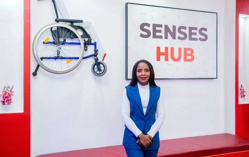 Woman in a blue vest smiling and standing in front of a sign that reads Senses Hub