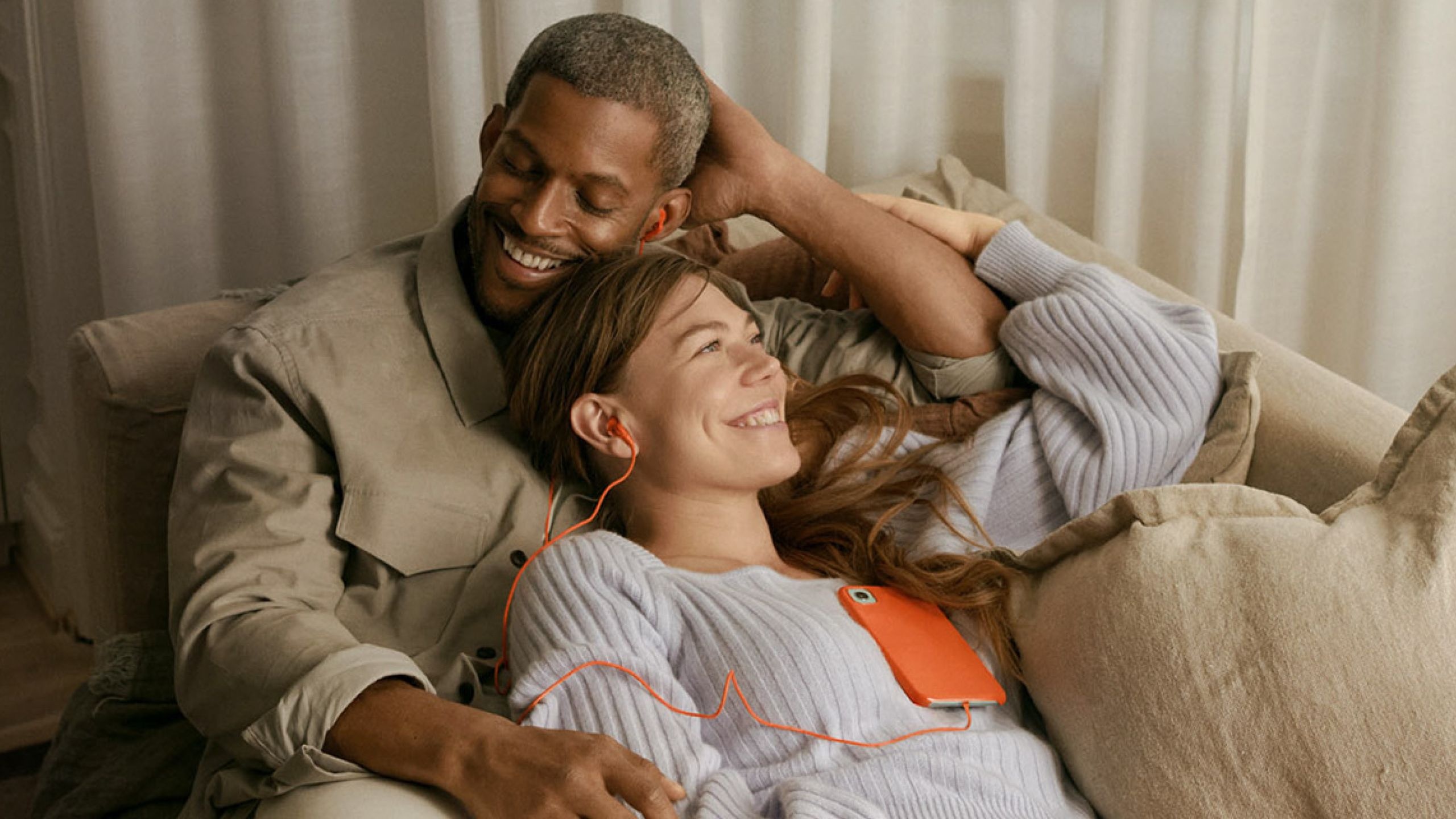 A happy couple lying on a sofa, listening to music with orange earphones and a smartphone.